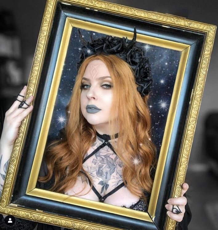 A woman in a picture frame. Long ginger hair, an Angel Rune on her forehead, wearing a crown of black roses.