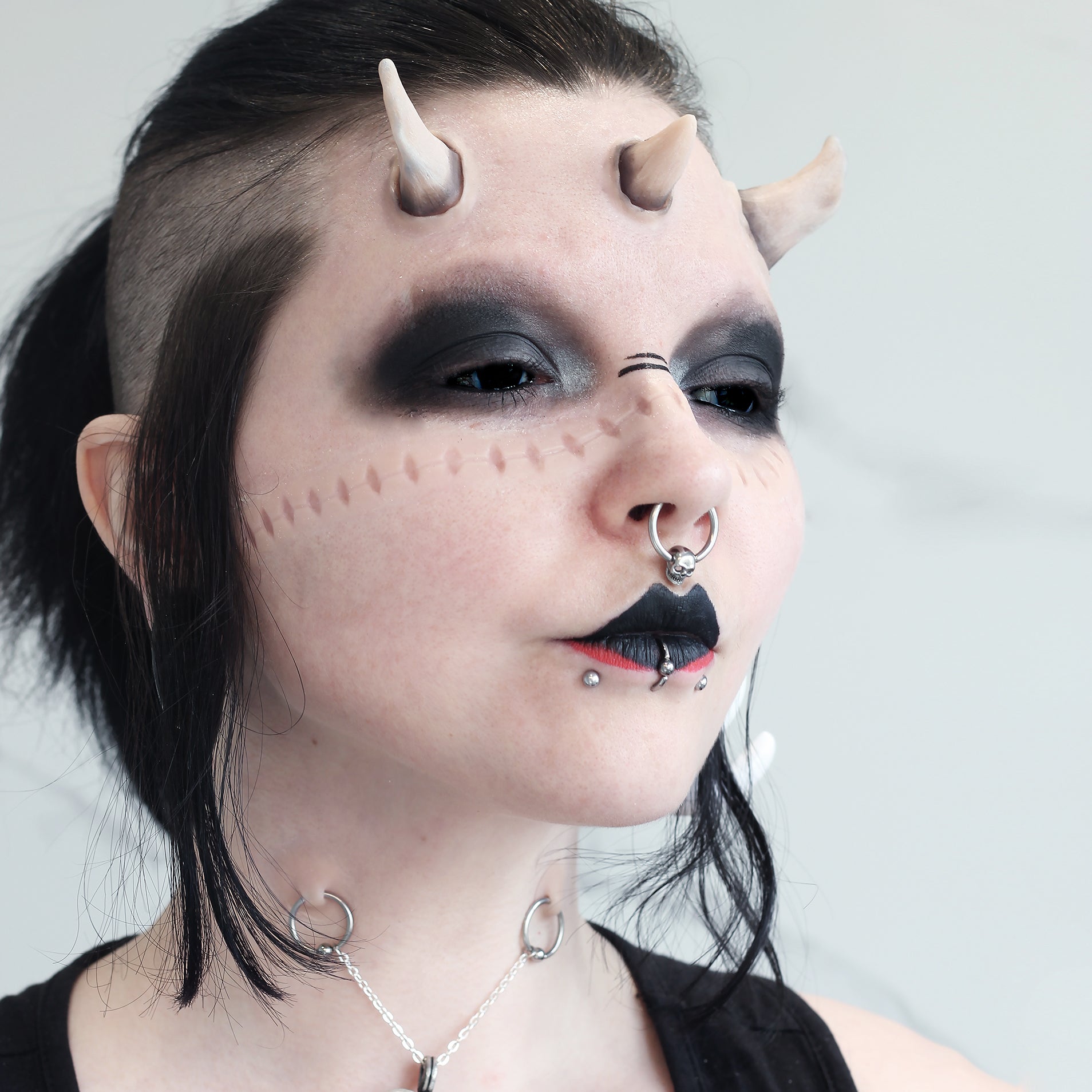 Woman with black hair wearing dark makeup, crooked horns, and scarification strip prosthetics