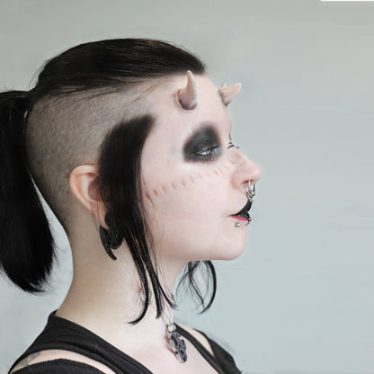 Woman with black hair wearing dark makeup, crooked horns and scarification strips prosthetics